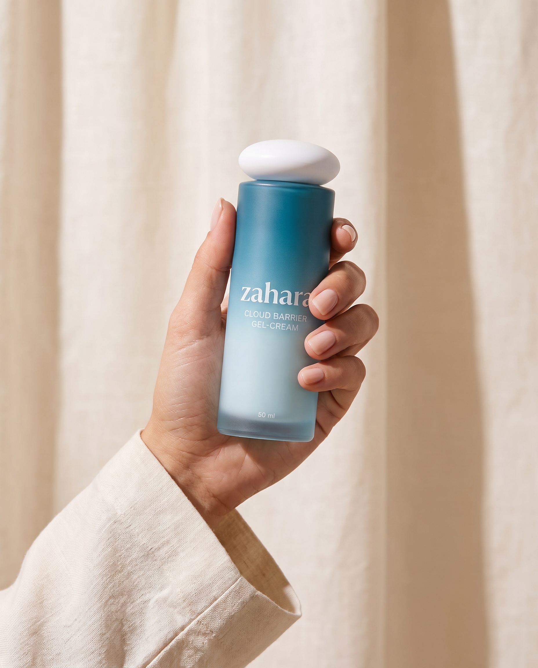 A hand in a cream linen sleeve holding the Zahara Cloud Barrier Gel-Cream bottle against a warm ivory linen backdrop.